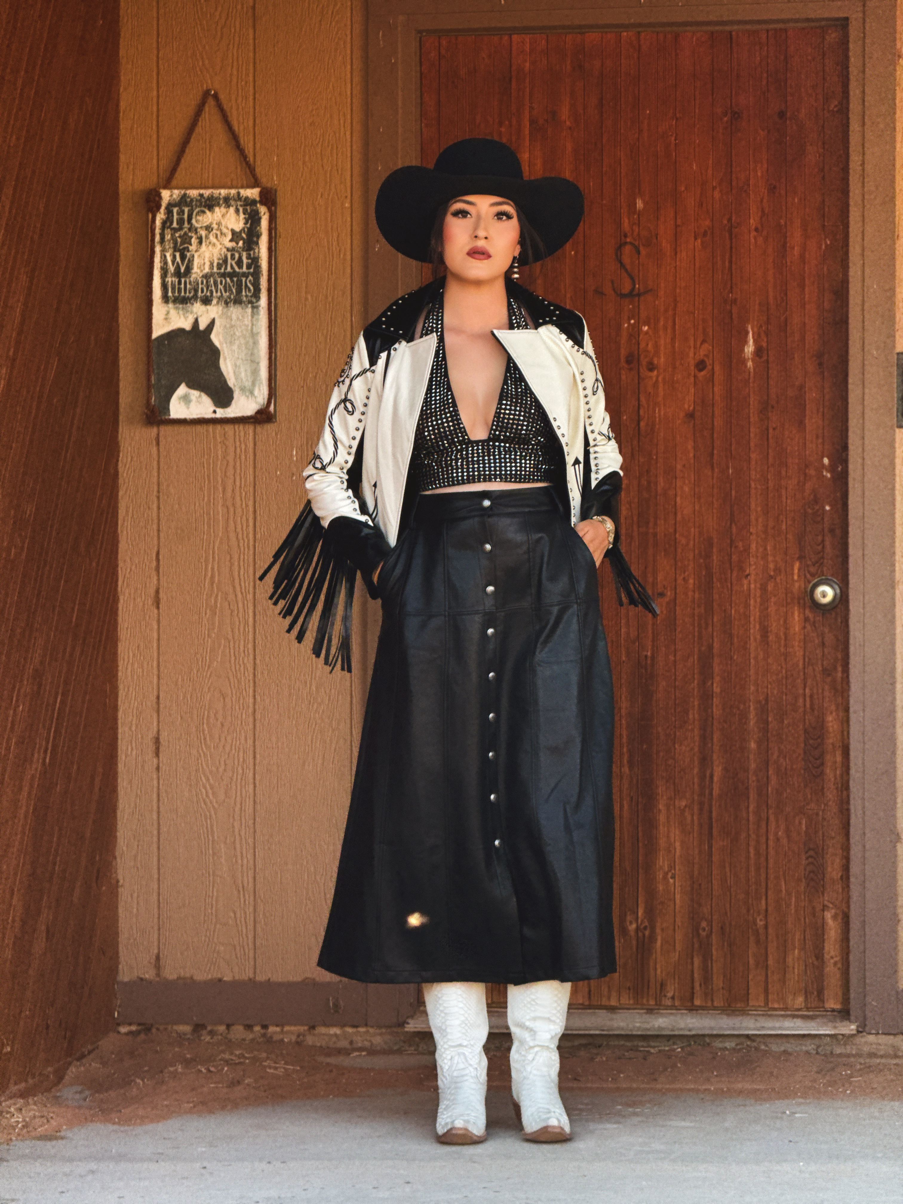 White Western fringe jacket with black studded accents, paired with a black embellished top, button-front leather-look midi skirt, cowboy hat, and white boots.
