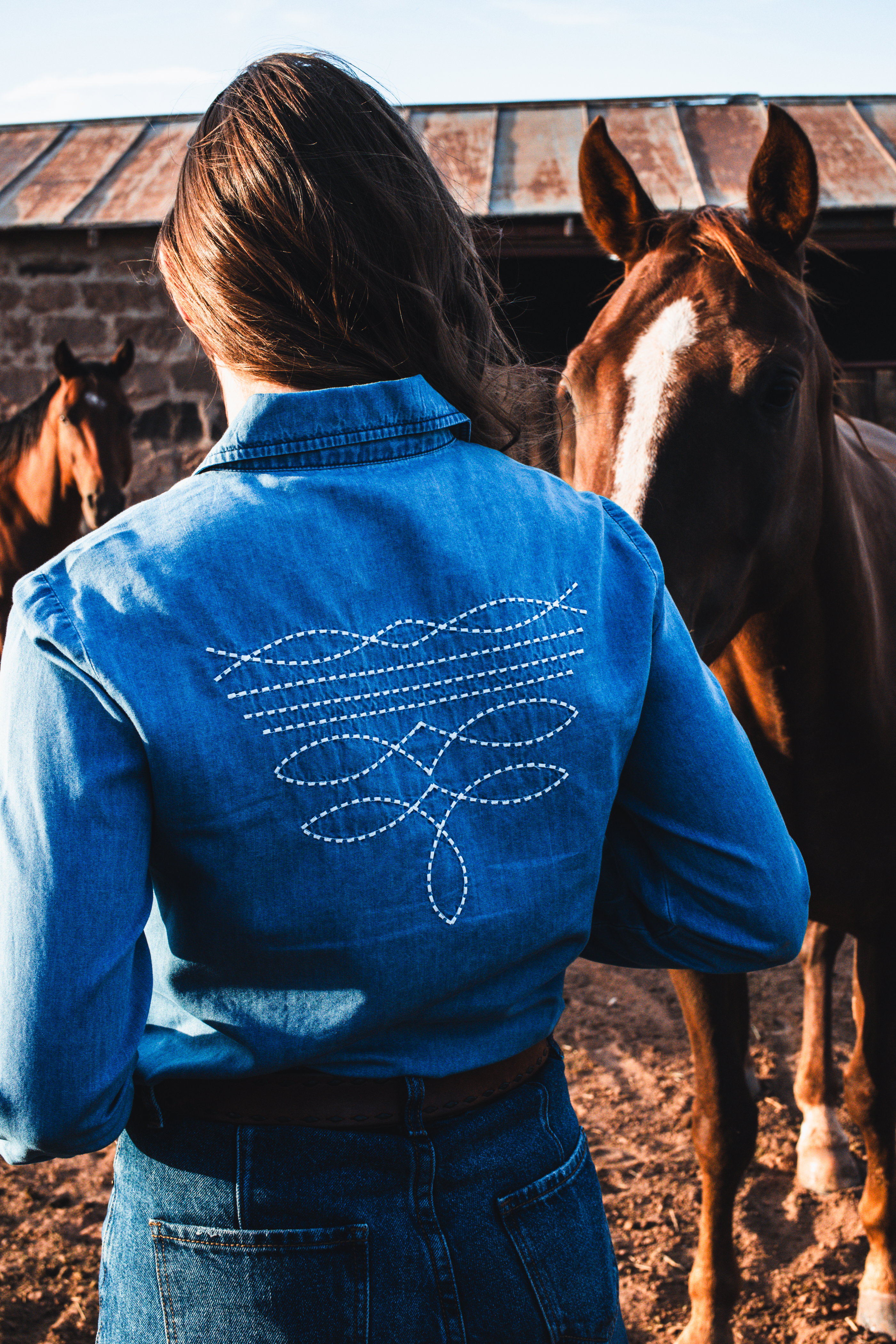 Cowgirl with a denin top with embroidered boot stitch on the back standing near horses