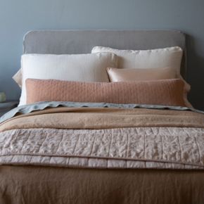 A cluster of pillows at the head of a monochromatic dressed bed with a body pillow spaning the width of the cluster