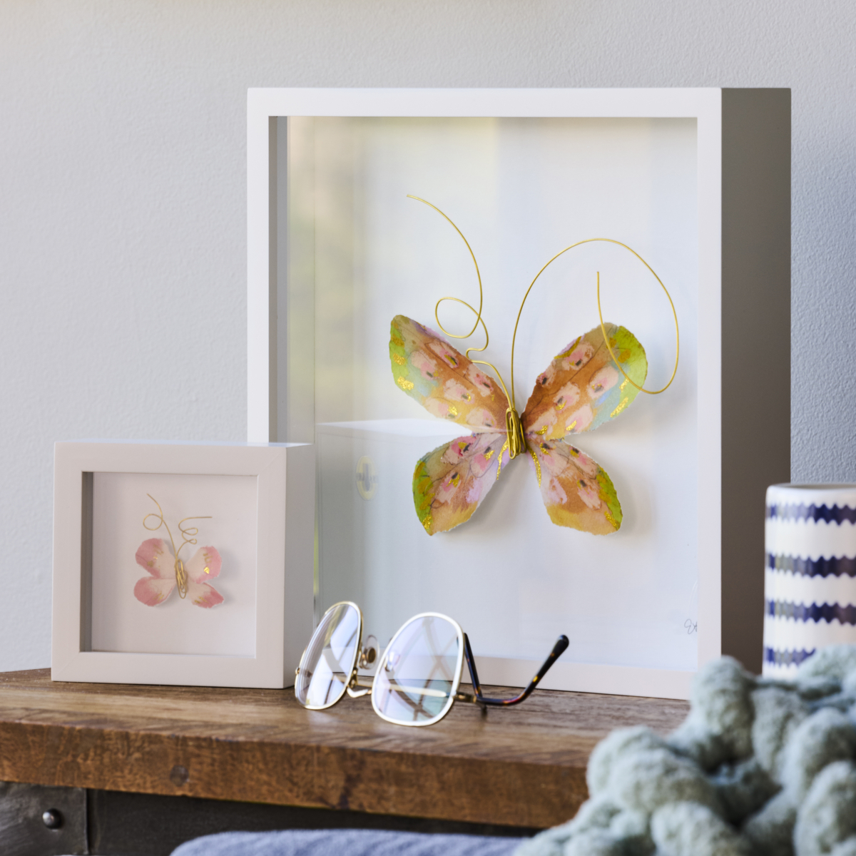 A butterfly wall art piece and butterfly shadow box sitting on a wood side table.