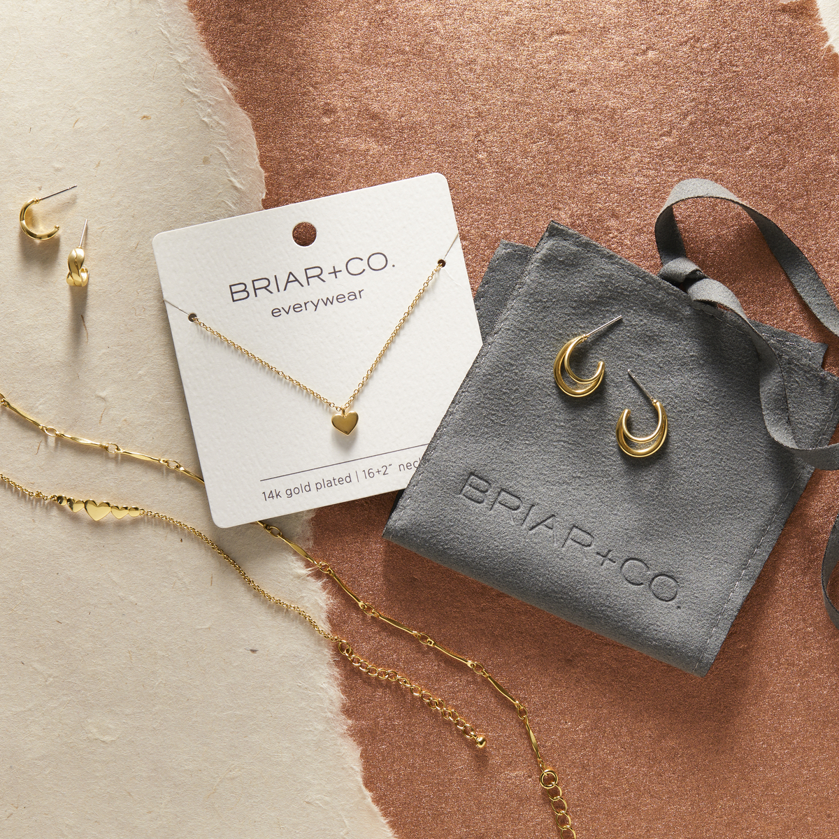 Gold jewelry displayed on a packaging backer card and with a small gray fabric gift bag on a brown and cream background