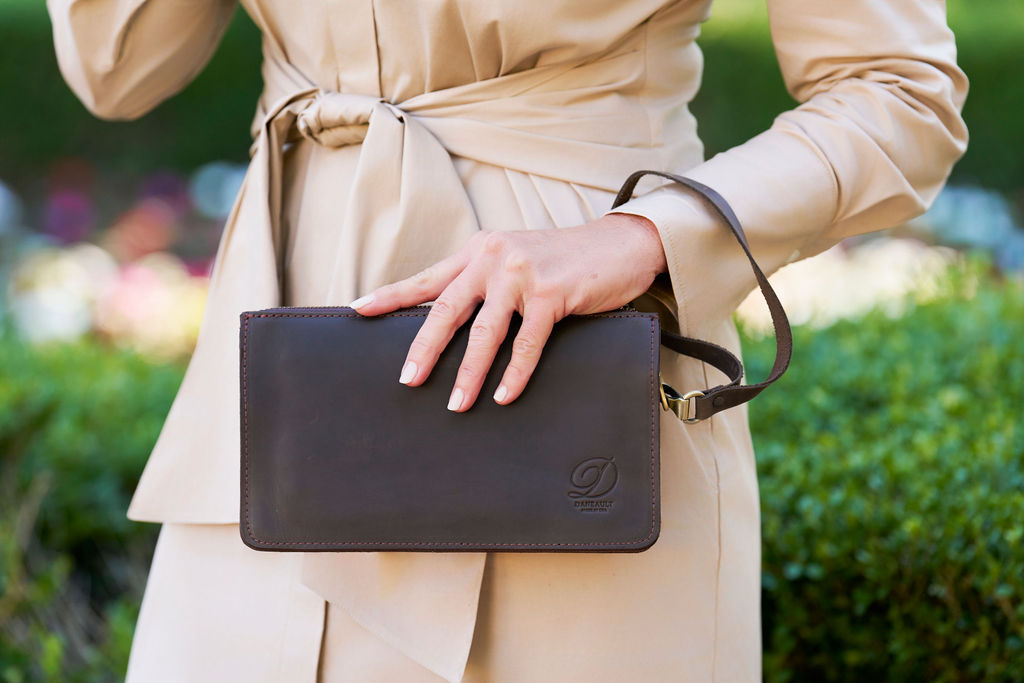 A woman wearing a beige trench coat holds a dark brown leather crossbody bag by Daneault with a subtle logo embossed on the front.